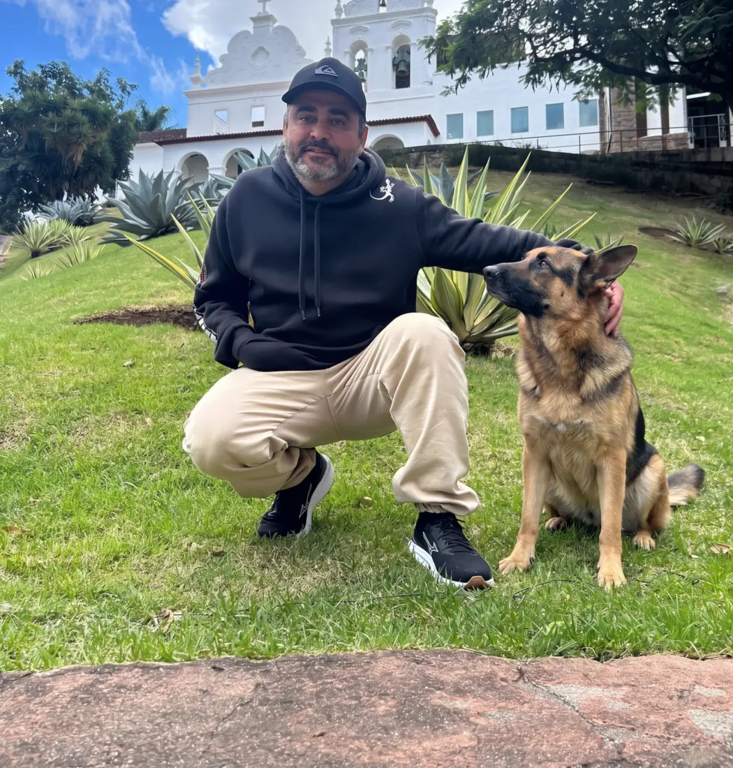 V. Júnior, adestrador de cães na Grande Vitória, agachado na grama ao lado de um pastor alemão adestrado
