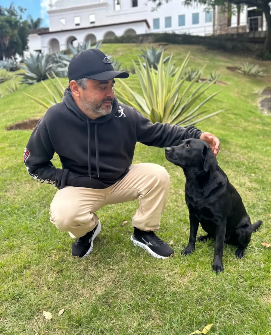V. Júnior agachado na grama dando comando de mão para um labrador preto sentado, atento